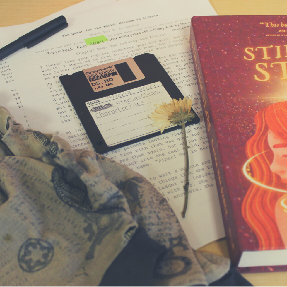 Photo of a copy of STILL THE STARS by Elayna Mae Darcy, which features a red cover and a girl with red hair who is surrounded by stars and light. Next to the book is a stack of papers, a black floppy disk, a pen, and a pair of writing gloves.
